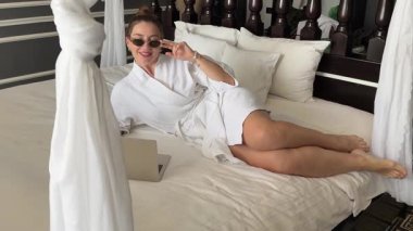 Yatakta dizüstü bilgisayarı olan siyah gözlüklü beyaz cüppeli bir kadın. Dans eden kız elini sallıyor