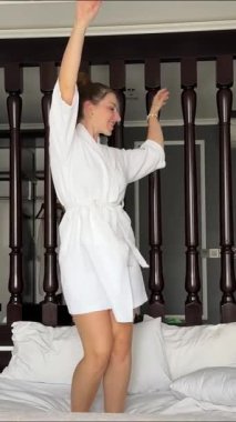 Kadın piyango talihlisi büyük zaferi kutluyor, dans ediyor, yatağa atlıyor, para saçıyor. Yavaş çekim videosu. Beyaz kadın cübbesi, yatakta dans eden kadın.