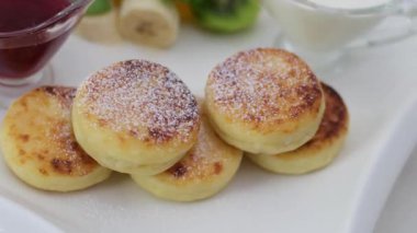 Reçelli şurup ve taze meyveli muzlu kivi ekşi krema ve beyaz tabakta ekşi kremayla servis edilen kızarmış süzme peynirli krep porsiyonlu yoğurt. Syrniki yapmak adım adım