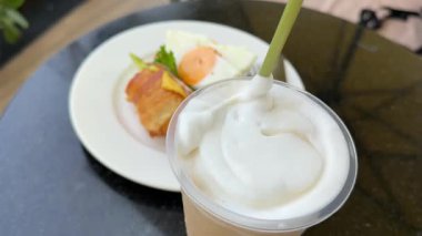 Kahve dükkanında kahve karıştırmak. Bol köpüklü kapuçino, buzlu latte kahveli kahve. Siyah bir masada yumurtalı kruvasan. Yüksek kalite 4k görüntü