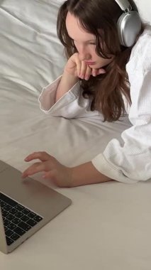Kulaklık takan kız çalışmaktan yoruluyor, dizüstü bilgisayarını bir kenara koyuyor ve gün içinde modern yatak odasında telefonuna sarılıyor. Yatakta beyaz bornozlu kız.