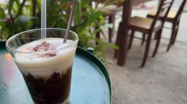 Kadın eli, tropik deniz manzaralı kafedeki ahşap masaya kokteyl ve metal pipetle birlikte uzun camlar koyuyor. Okyanuslu çatı restoranındaki bardak altlığına buzlu kahve ya da smoothie koymak.