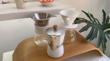 Kahve dükkanındaki masada çeşitli türde kahve makineleri var, bir filtre kahve makinesi ve diğer türleri, bir palmiye yaprağının arka planında, hepsi beyaz tonlarda. Yüksek kalite 4k görüntü