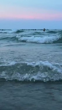  Deniz dalgaları kıyıya vuruyor ve yavaş çekim kamerasından yükseliyor. İnsanlar arka planda akşam güneşinde yüzüyor. Dağlarda deniz sahillerinde dinleniyor. Yüksek kalite 4k 