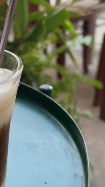 Kadın eli, tropik deniz manzaralı kafedeki ahşap masaya kokteyl ve metal pipetle birlikte uzun camlar koyuyor. Okyanuslu çatı restoranındaki bardak altlığına buzlu kahve ya da smoothie koymak.