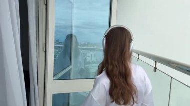 Genç, kıvırcık saçlı, kulaklık takan bir kadın şehre tepeden bakan bir terasta dans ediyor. Kadın hayattan, temiz havadan ve günbatımından zevk alıyor. Yüksek kalite 4k görüntü