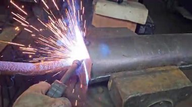 Elektrikli metal kaynak atölyesinde çalışan genç bir zanaatkar için yavaş çekim, kıvılcım saçan bir ışık ve sıcak termal duman.