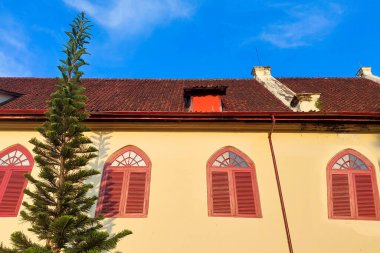 Kemerli kırmızı pencereli, sarı cepheli ve parlak mavi gökyüzünün altındaki eski kiremitli çatılı tarihi bir binanın yakın görüntüsü..