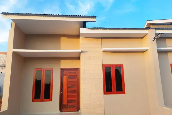 Modern minimalist tasarımı olan basit bir ev, bej duvarlar, ahşap kapılar, ve kırmızı çerçeveli pencereler, açık mavi bir gökyüzünün altında, önden görülüyor..