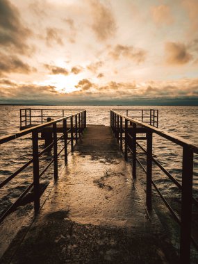 Ufkun sükunetle buluştuğu yerde, metal bir rıhtım günbatımından öpülen denizin boyalı dinginliğine uzanır. Altın Saat.