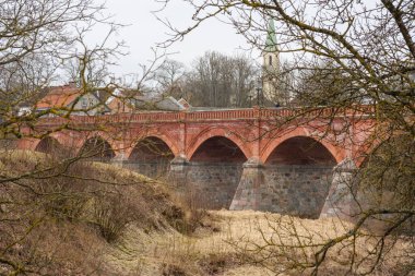 Venta Nehri 'nin sakin sularında Kuldiga' nın ikonik kırmızı tuğla köprüsünden geçmek, her adımın zamanda bir yolculuk olduğu yerden