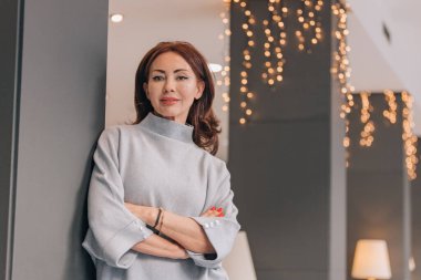 Kollarını kavuşturup kollarına yaslanmış şık bir kadın Noel ışıklarıyla kameraya bakıyor..