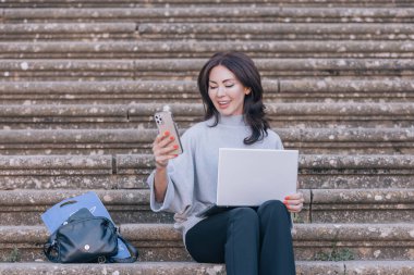 Güzel ve zarif bir kadın eski merdivenlerde oturmuş gülümsüyor ve telefonuna bacaklarında bilgisayarla bakıyor..