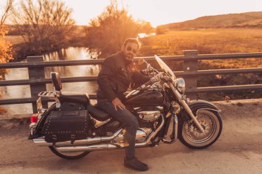 Motosikletli, sakallı ve güneş gözlüklü sonbahar günbatımında nehrin kenarında kameraya bakan.