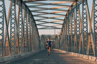 Genç sporcu, gün batımında eski bir metal köprüde gülümsüyor. Spor konsepti.