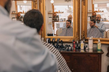 Bir adam berber dükkanında saçını kestiriyor. Berber adamın saçını keserken yanındaki sandalyede başka bir adam oturuyor. Berber adamın saçını kesmek için bir makas kullanıyor.