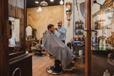 Barber cuts the hair of a young mans head with scissors and comb on a beautiful antique iron chair in a barber shop.