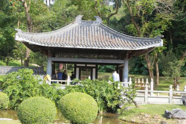 Bir grup insan parktaki bir pagoda 'nın dışında duruyor. Pagoda çalılar ve ağaçlarla çevrili ve yakınlarda bir gölet var. Atmosfer huzurlu ve huzurlu.