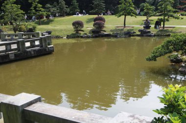 Üzerinde köprü olan bir gölet ve köprüde birkaç kişi oturuyor. Su yeşil ve gökyüzü mavi.