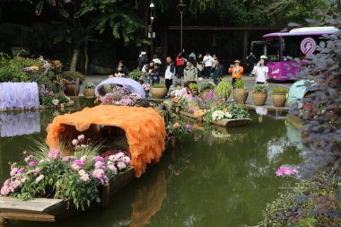 Bir grup insan arka planda mor bir otobüsle bir göletin etrafında duruyorlar. Gölet tekneler ve çiçeklerle dolu. Huzurlu ve huzurlu bir ortam yaratıyor.