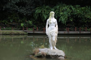 Göletin önündeki bir kayanın üzerinde duran bir kadın heykeli. Heykel beyazdır ve yeşilimsi bir tonu vardır. Gölet ağaçlar ve çalılarla çevrili. Huzurlu ve huzurlu bir atmosfer yaratıyor.