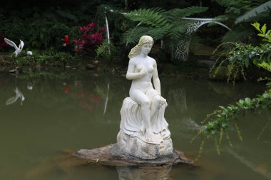 Göletteki bir kayanın üzerinde bir kadın heykeli oturuyor. Heykel beyaz ve gölet yeşil.