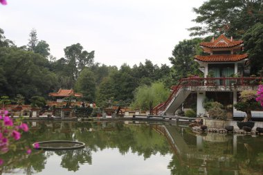 Arkasında köprü ve bina olan bir gölet. Gölet ağaçlar ve çalılarla çevrili.
