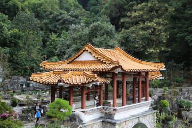 Beyaz çatılı ve ahşaptan yapılmış küçük bir bina. Çatı eğimli ve kırmızı bir rengi var. Binanın yakınında yürüyen iki kişi var.