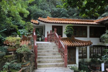 Kırmızı çatılı ve beyaz süslemeli bir ev. Evin beyaz bir verandası ve kırmızı bir merdiveni var. Ev ağaçlar ve bitkilerle çevrili.