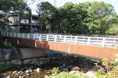 Arka planda yeşil bir ağaç olan nehir üzerinde bir köprü. Köprü tahtadan yapılmıştır ve beyaz bir parmaklığı vardır.