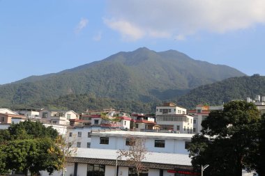 Bir dağ sırası bir şehrin arka planında görülebilir. Şehir, bazıları uzun ve beyaz olmak üzere binalar ve evlerle dolu. Gökyüzü açık ve mavidir ve güneş ışıl ışıl parlar.
