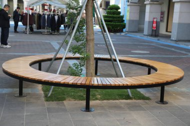 Ortasında ağaç olan yuvarlak ahşap bir bank. Bankın etrafı tuğla kaldırımlarla çevrili.