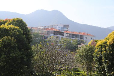 Bir yerleşim alanının arka planında bir dağ sırası vardır. Evler beyaz ve kırmızı ve bölgede birçok ağaç var.