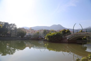 Bir köprü ortasında yeşil bir köprü olan bir nehri kapsar. Köprü ağaçlarla çevrili ve arka planda bir dağ var.