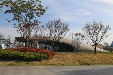 Kavisli çatılı büyük bir bina bir parkın önünde duruyor. Park ağaçlar ve çalılarla doludur ve çimenler kuru ve kahverengidir. Bina halka açık bir yer gibi görünüyor.