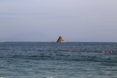 Kayalık bir ada, ön planda büyük bir su kütlesiyle uzaktan görülebilir. Gökyüzü bulutlu, sahneye kasvetli bir hava katıyor.
