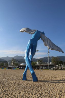 Sahile yayılmış kanatları olan büyük mavi bir kuş heykeli. Heykel metalden yapılmış ve kumun üzerinde duruyor. Kumsal boş ve gökyüzü açık ve mavi.