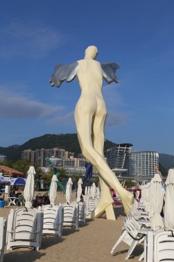 Kanatlı bir adamın büyük bir heykeli plajda beyaz sandalyelerin yanında duruyor.