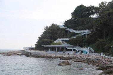 Büyük bir kaya duvarı ve merdiveni olan bir sahil. Kumsal boş ve gökyüzü bulutlu.