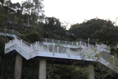 Tepenin üstündeki köprüde beyaz bir merdiven var. Köprü ağaçlarla çevrili ve gökyüzü manzaralı.