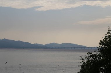 Bir köprü, arka planda bir dağ sırası olan bir su kütlesini kapsar. Gökyüzü bulutlu, karamsar bir atmosfer yaratıyor