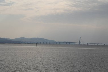 Köprü, arka planda bir dağ bulunan bir su kütlesini kapsar. Gökyüzü bulutlu ve su sakin.