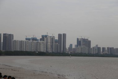 Arka planda büyük bir su kütlesi olan bir şehir silueti. Gökyüzü bulutlu ve su sakin.