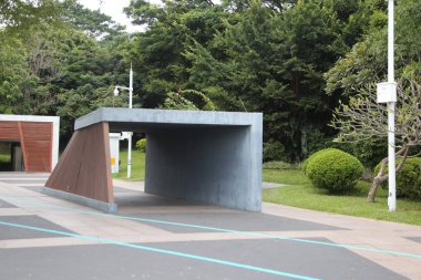 Yeşil çatılı büyük bir beton yapı yeşil bir parkın önünde duruyor. Bina ağaçlar ve çalılarla çevrili ve bölgede birkaç saksı bitkisi var. Huzurlu bir sahne.