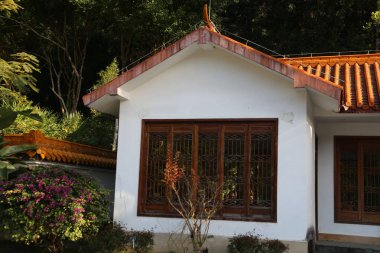 Kırmızı çatılı ve kahverengi panjurlu beyaz bir ev. Panjurlar açık, içeri güneş ışığı giriyor. Ev çiçekli ve çalılı bir bahçeyle çevrili.