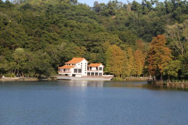 Arka planda ağaçlar olan bir göl üzerinde bir ev oturur. Ev beyaz ve kırmızı bir çatısı var. Göl sakin ve huzur dolu, ev ona bakıyor. Sahne huzurlu ve resimli.