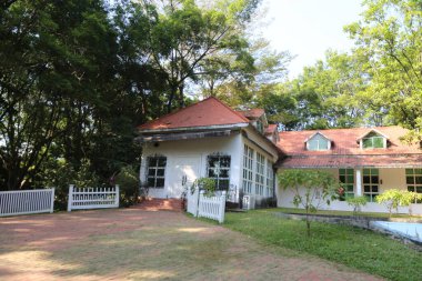 Beyaz çatılı ve beyaz çitli büyük bir ev. Ev ağaçlarla çevrili ve yemyeşil bir çimenliği var.