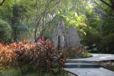 Bir taş heykel yemyeşil bir bahçenin ortasındadır. Heykel, kırmızı yapraklı büyük bir çalılık da dahil olmak üzere çeşitli bitkilerle çevrilidir. Bahçe bakımlı görünüyor.