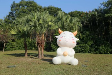 Beyaz doldurulmuş bir hayvan ineği çimenli bir tarlada oturuyor. İnek ağzında sarı bir nesne tutuyor. Sahne huzurlu ve huzurlu, inek mutlu ve rahat görünüyor.