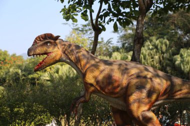 Parkta bir dinozor heykeli duruyor. Heykel kahverengidir ve büyük bir kafası vardır. Heykel ağaçlar ve çalılarla çevrili.